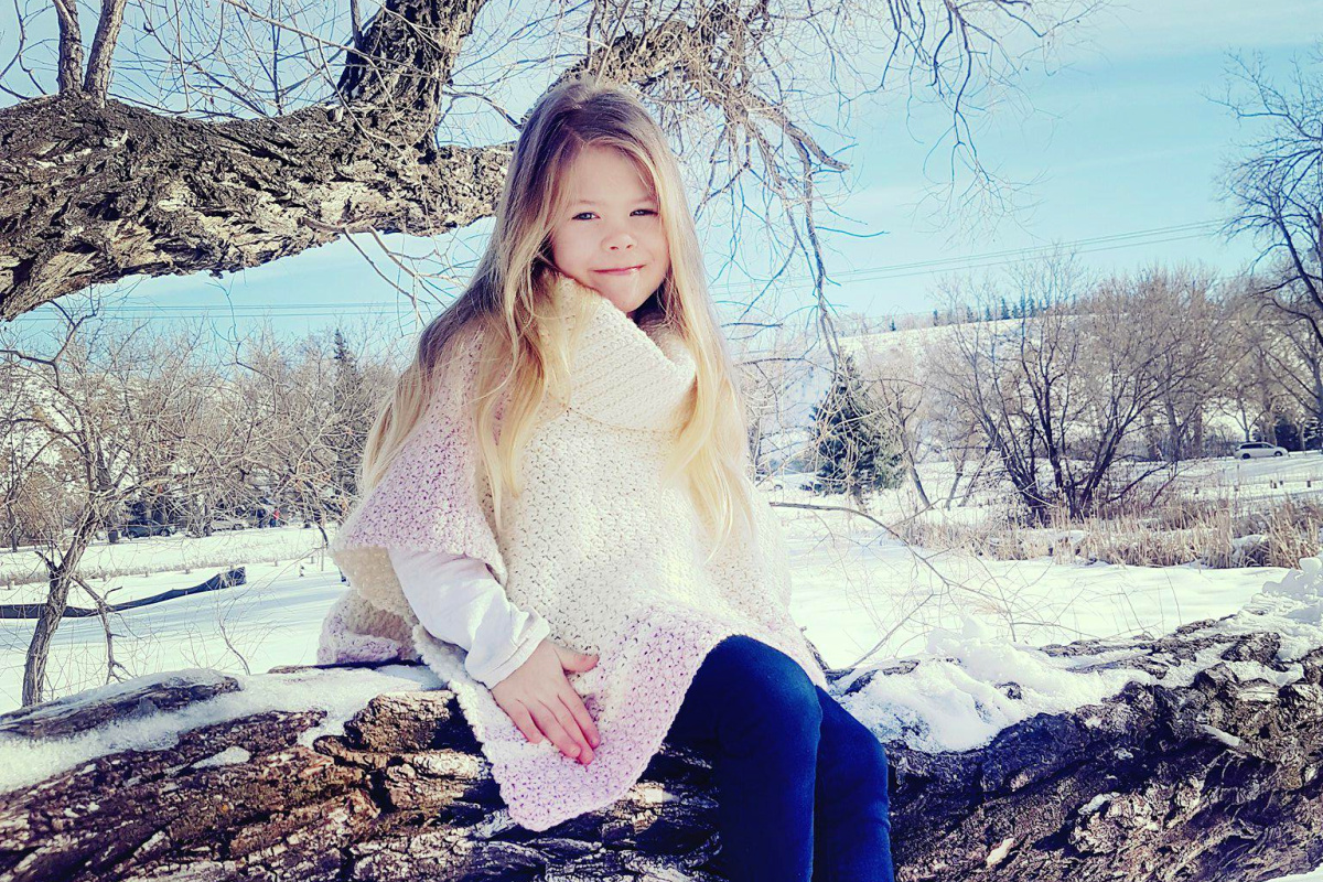 A child wearing a cream and pink colored poncho sitting on a log with a snowy background.