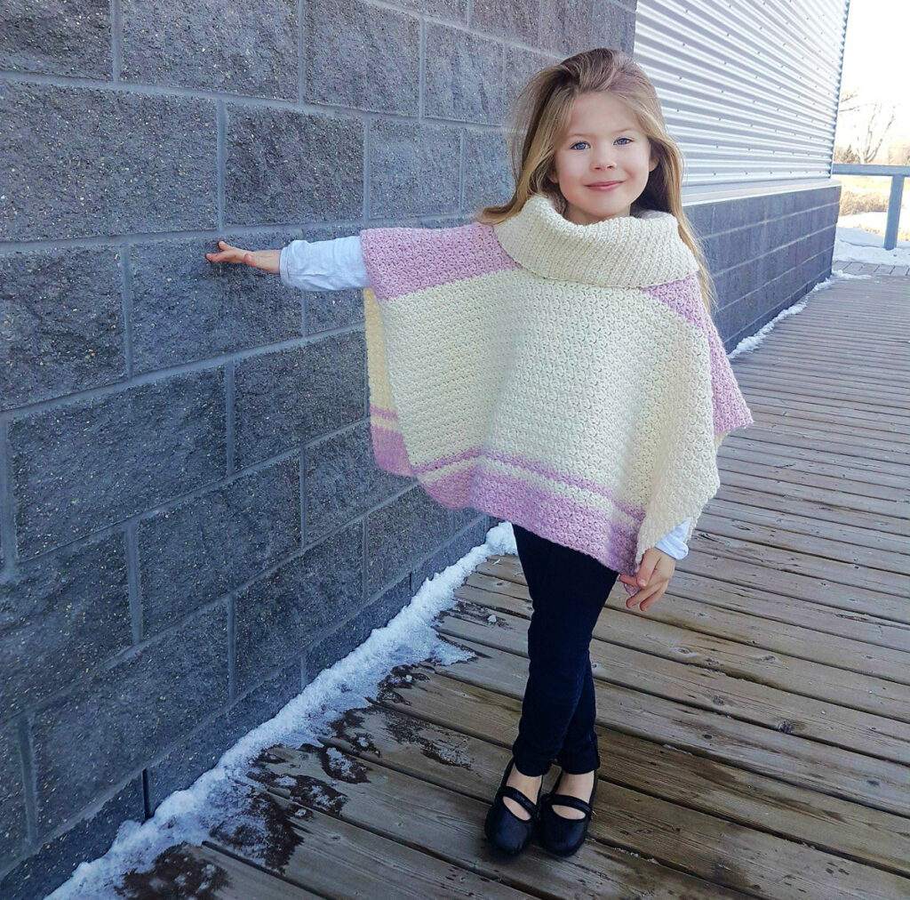A child wearing a cream and pink colored poncho and black bottoms standing with her hand on the wall.