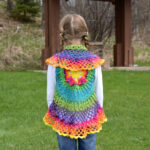 Girl wearing a colorful mandala crochet vest and a white shirt standing outside with her back to the camera.