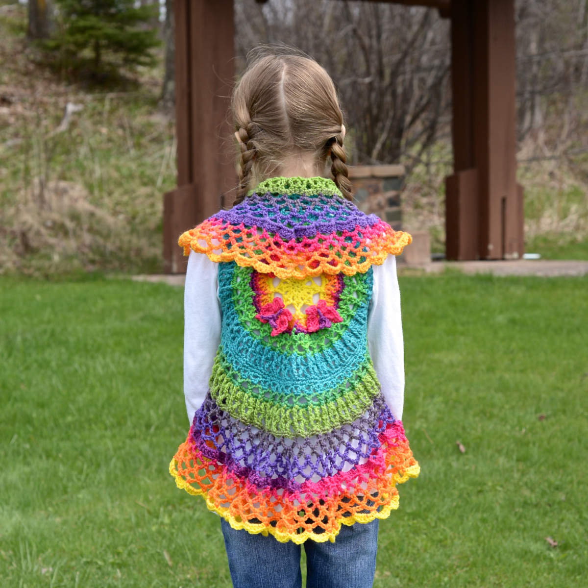 Girl wearing a colorful mandala crochet vest and a white shirt standing outside with her back to the camera.