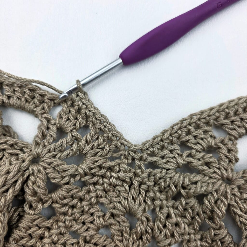 A crochet hook working the edging of a crochet granny square bag.