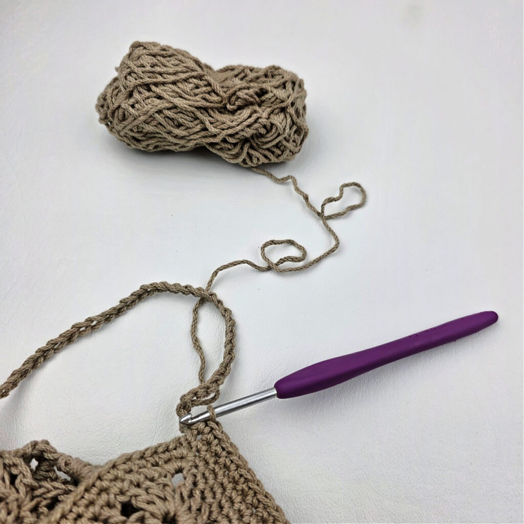 A crochet hook working the strap of a crochet granny square bag.