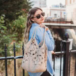 A girl wearing sunglasses and a blue shirt holding a crochet tote bag over her shoulder next to a fence.