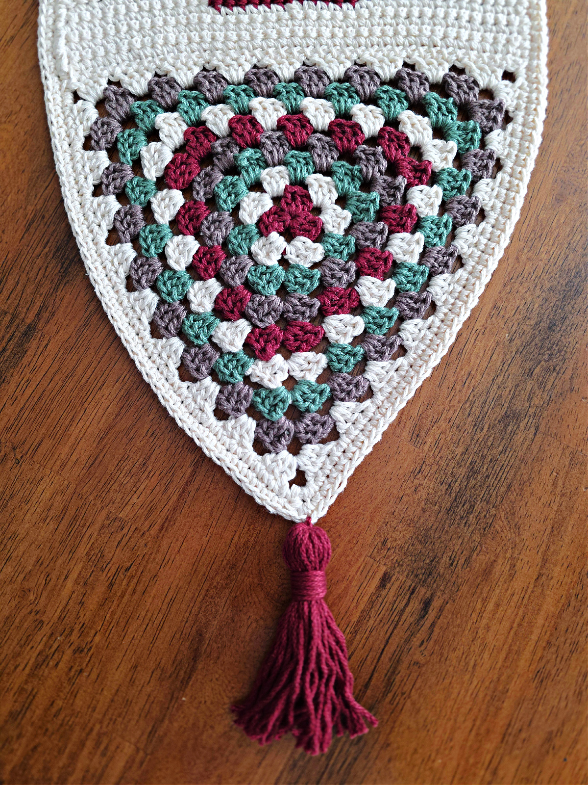 A crochet granny triangle made from brown, red, green, and white yarn with a tassel.