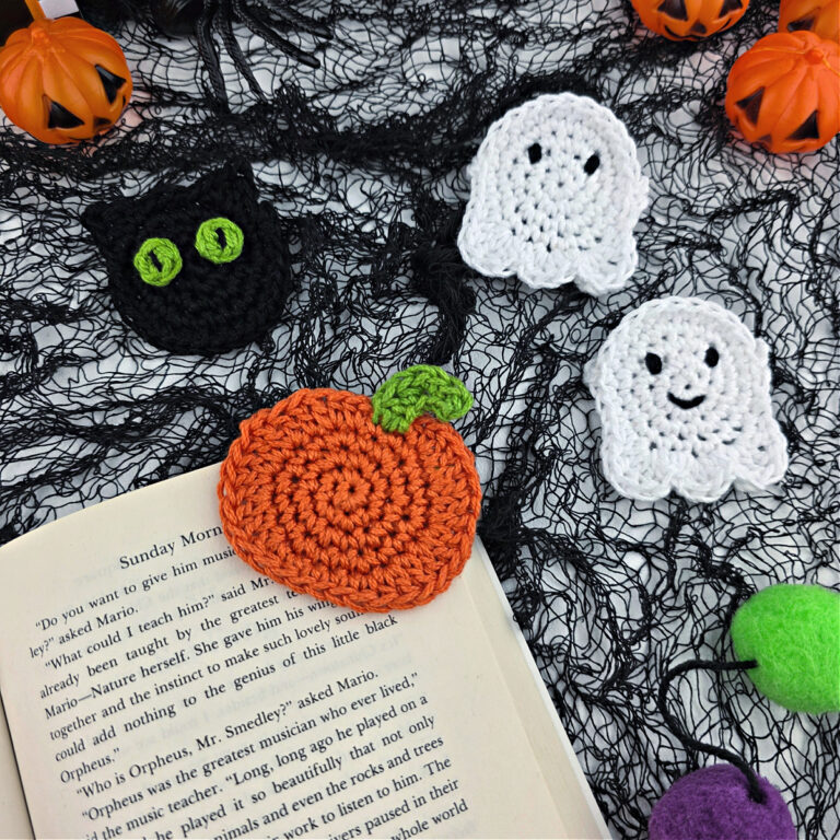 A crochet pumpkin bookmark attached to a book with a crochet ghost and cat in the background.