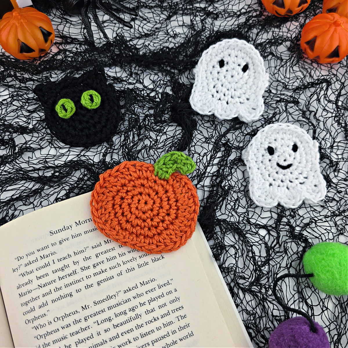 A crochet pumpkin bookmark attached to a book with a crochet ghost and cat in the background.