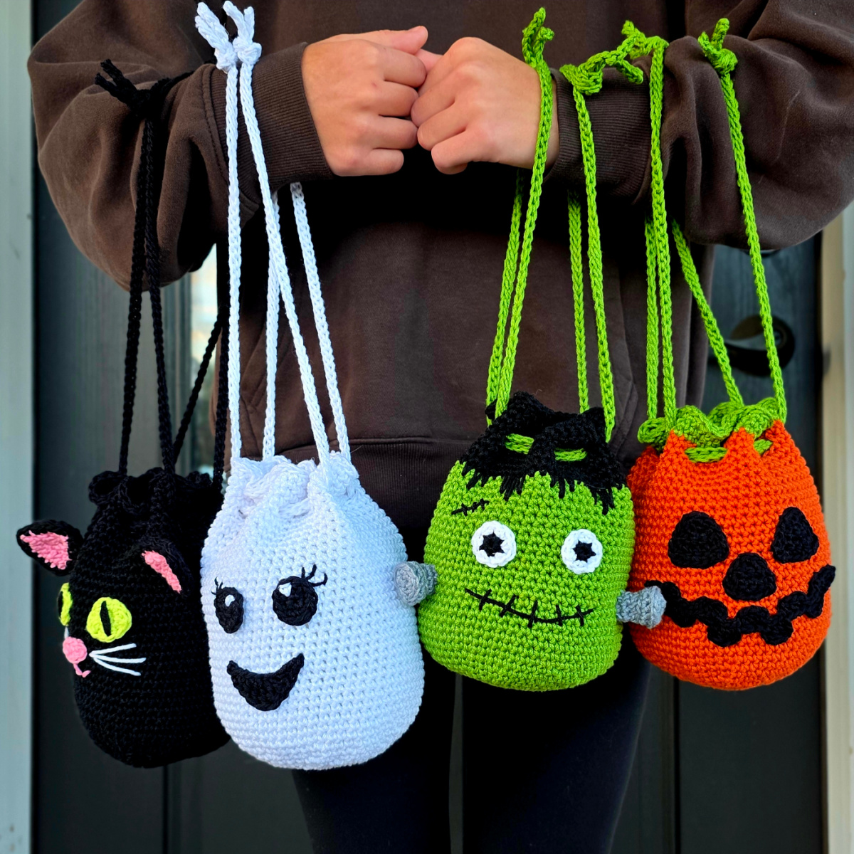 A person holding monster, jack-o-lantern, ghost, and black cat drawstring treat bags on their arms.