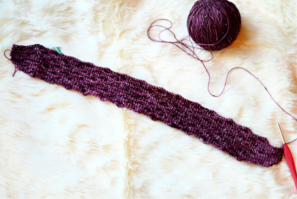 A purple crochet stitch with a red crochet hook and purple ball of yarn against a white background.