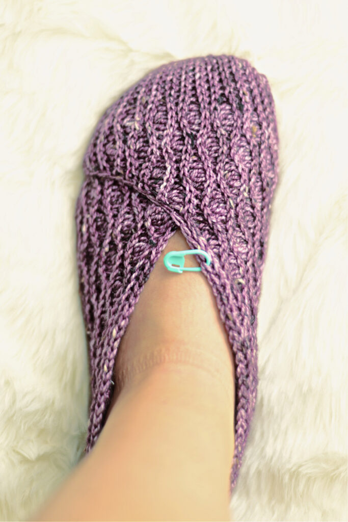 A purple crochet kimono slipper on feet with a stitch marker against a white background.