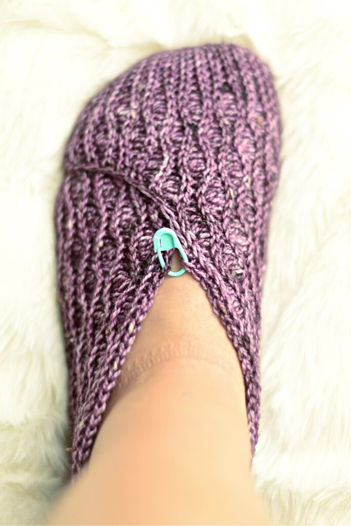 A purple crochet kimono slipper on feet with a stitch marker against a white background.
