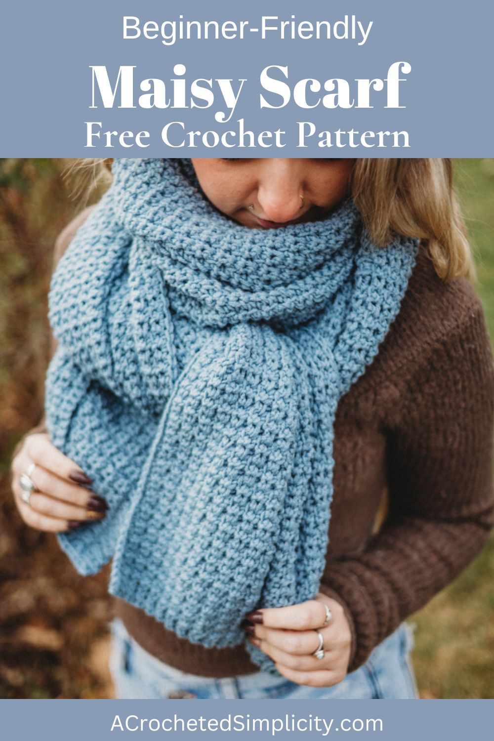 Woman wearing a brown sweater with a large blue crochet scarf wrapped around her neck, holding the edges, standing in a yard.