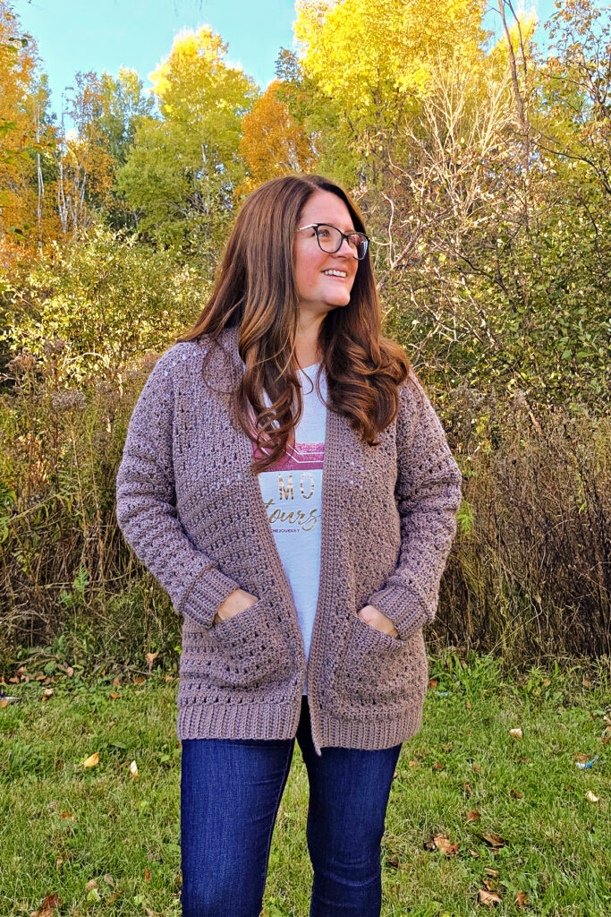 A woman wearing a brown crochet cardigan sweater and blue jeans with her hands tucked into the pockets.