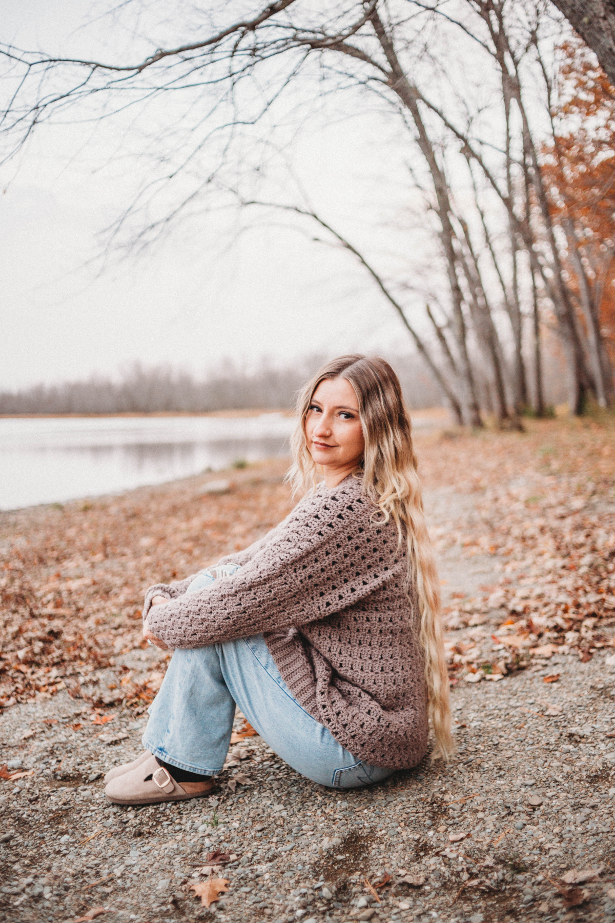 A woman with blonde hair wearing a brown crochet cardigan sweater sitting on the ground in front of the water.
