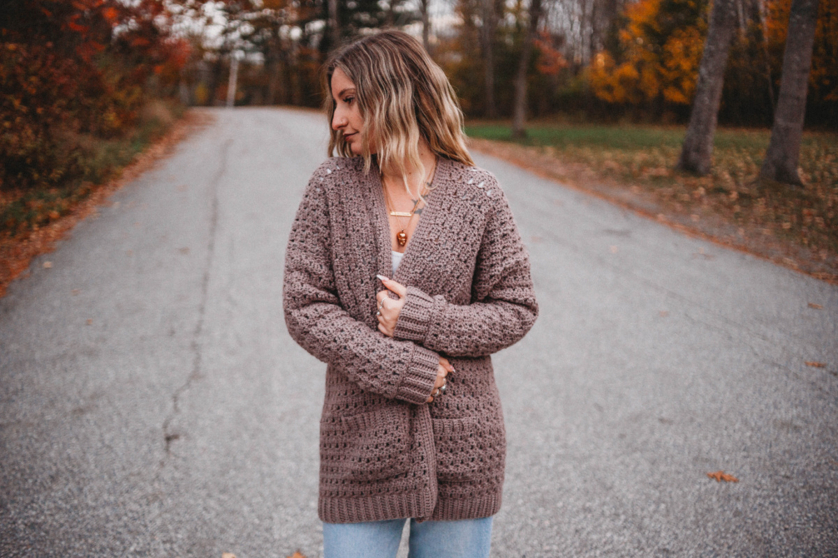 A woman with blonde hair wearing a brown crochet cardigan sweater holding the sides together.