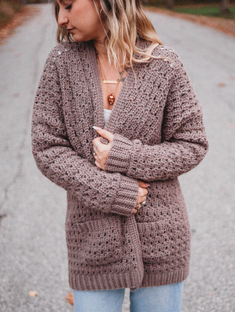 A woman with blonde hair wearing a brown crochet cardigan sweater holding the sides together.