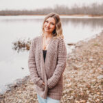 A woman with blonde hair wearing a brown crochet cardigan sweater and faded blue jeans standing in front of the water.