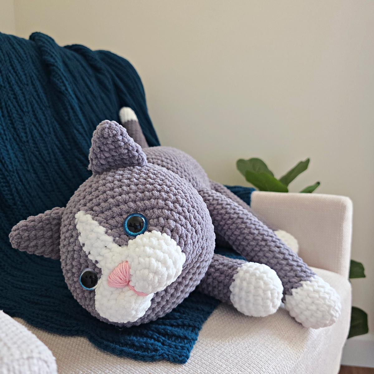 A large gray amigurumi cat on a blue blanket spread on a sofa with a plant next to it.