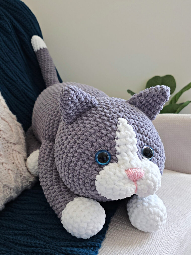 A large gray amigurumi cat on a blue blanket spread on a sofa with a plant next to it.