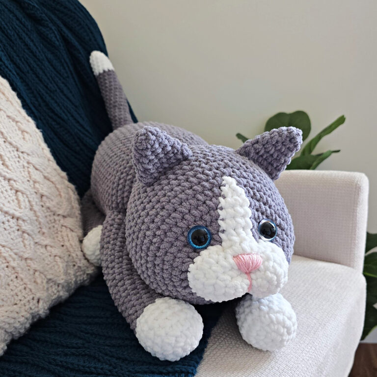 A large gray amigurumi cat on a blue blanket spread on a sofa with a plant next to it.