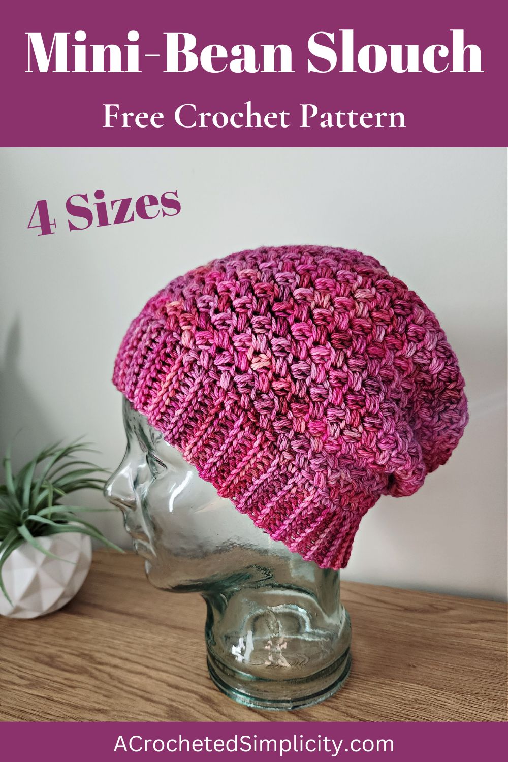A pink crochet beanie on a glass mannequin head on a wooden shelf with a plant next to it.
