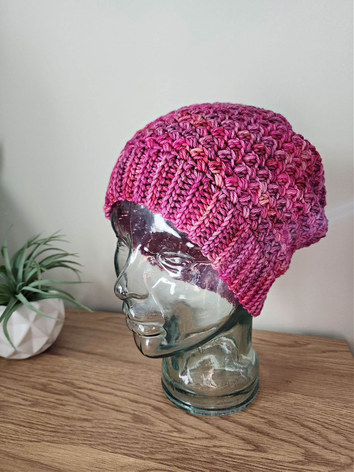 A pink crochet beanie on a glass mannequin head on a wooden shelf with a plant next to it.
