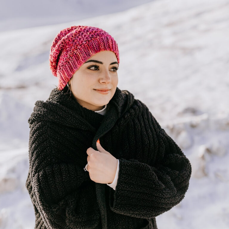 Girl wearing a pink crochet beanie and black coat with the snow behind her.