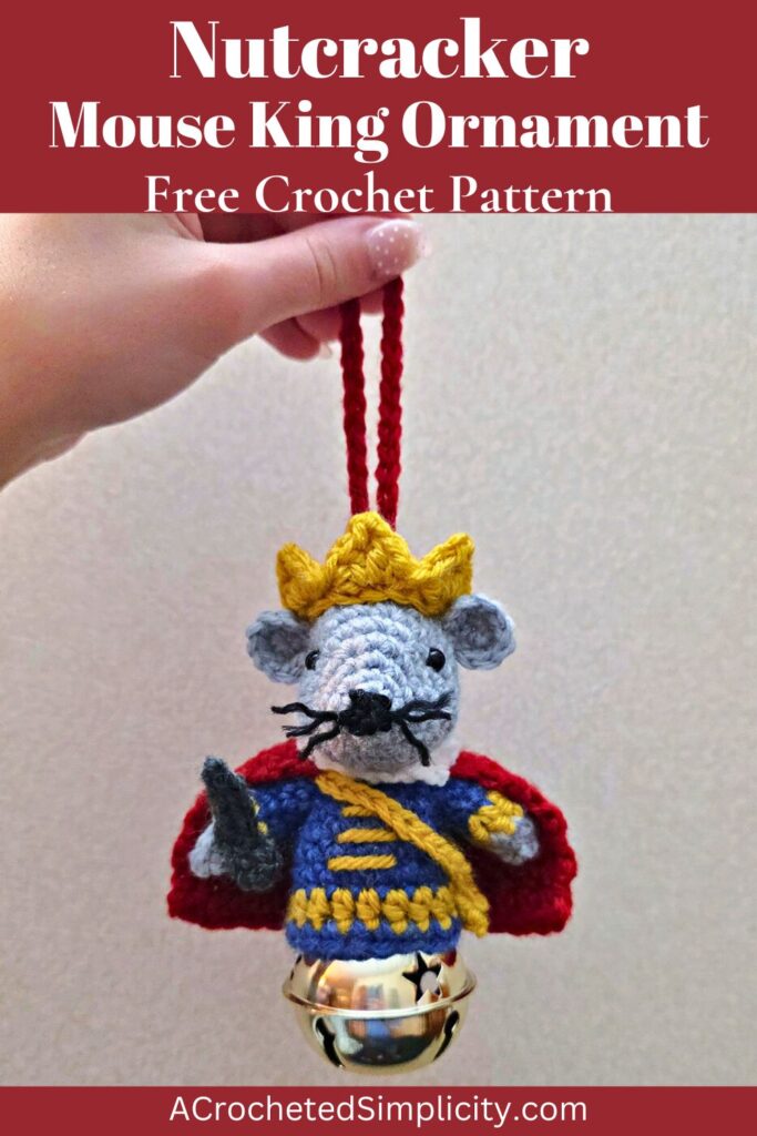 A cute Nutcracker Mouse King crochet ornament being held in front of a wall by a hand with cute painted nails.