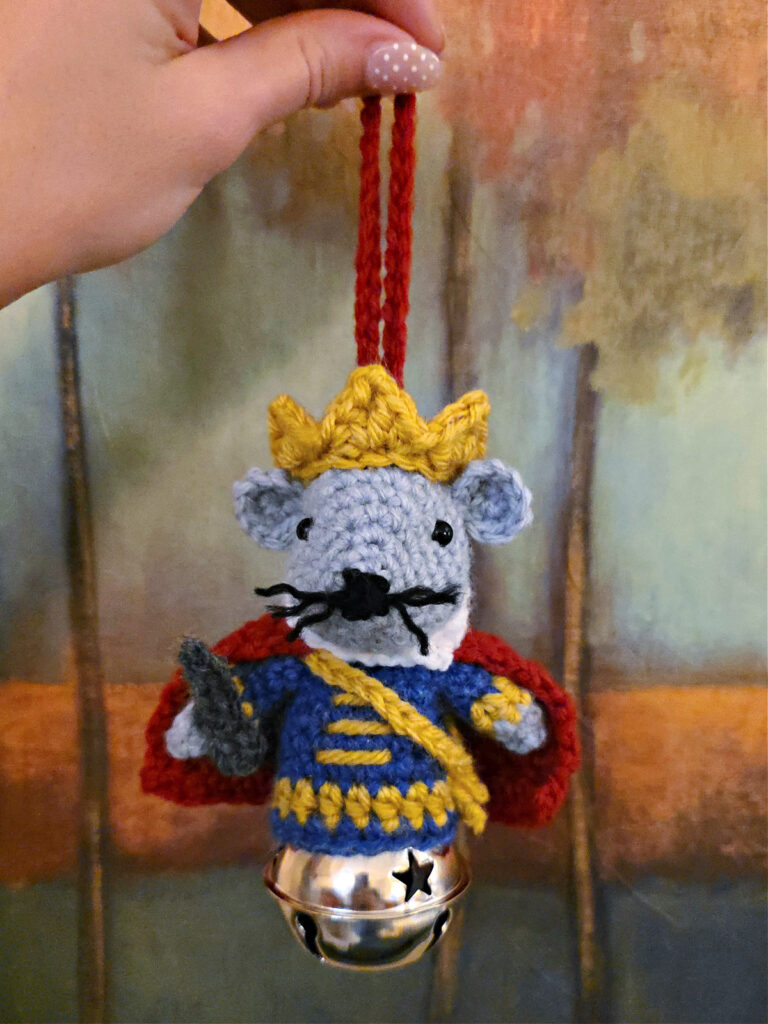 Nutcracker Mouse King crochet ornament being held in front of a painting in the woods with colorful leaves.