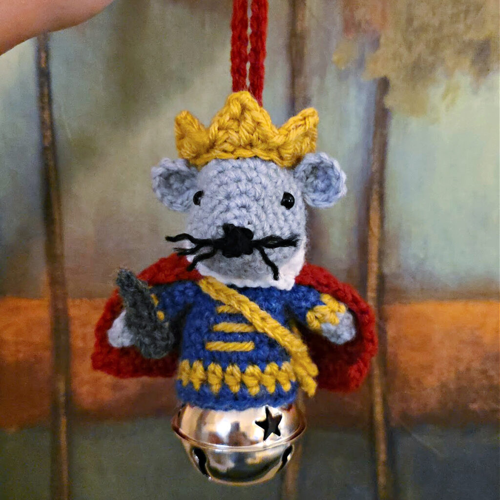 Nutcracker Mouse King crochet ornament being held in front of a painting in the woods.