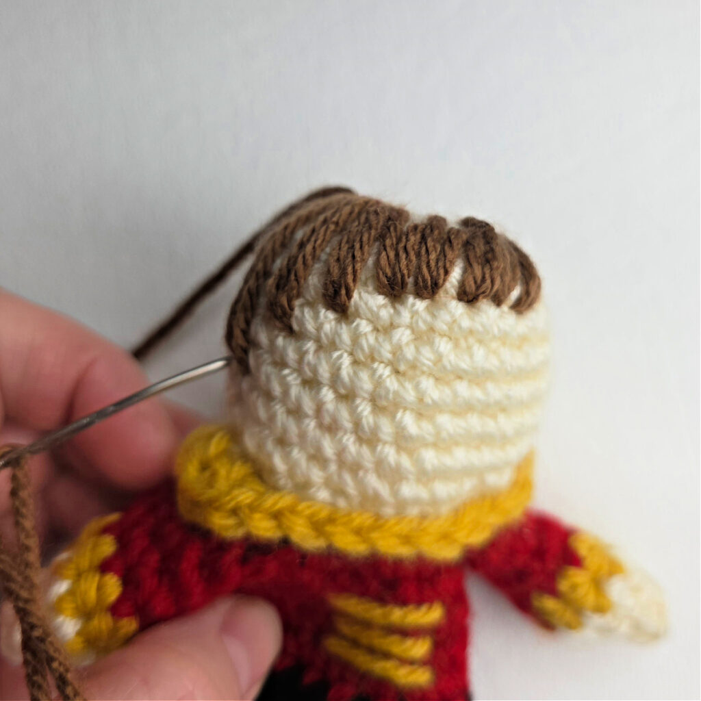 A yarn needle and brown yarn being stitched on the head of a crochet ornament.