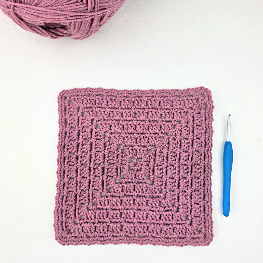 A pink block stitch granny square with a ball of yarn in the corner and a blue crochet hook.