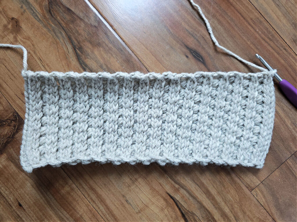 A grey crochet strip with a purple crochet hook against a wooden floor.