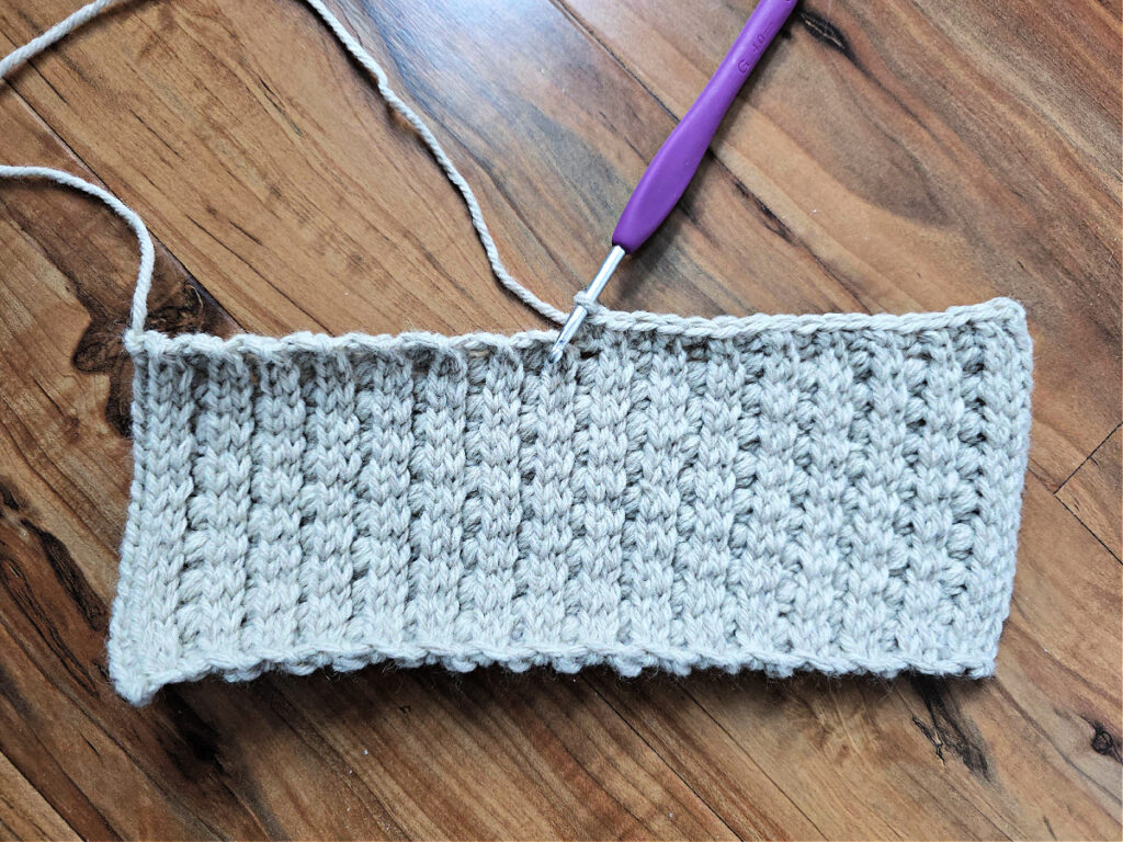 A grey crochet strip with a purple crochet hook against a wooden floor.