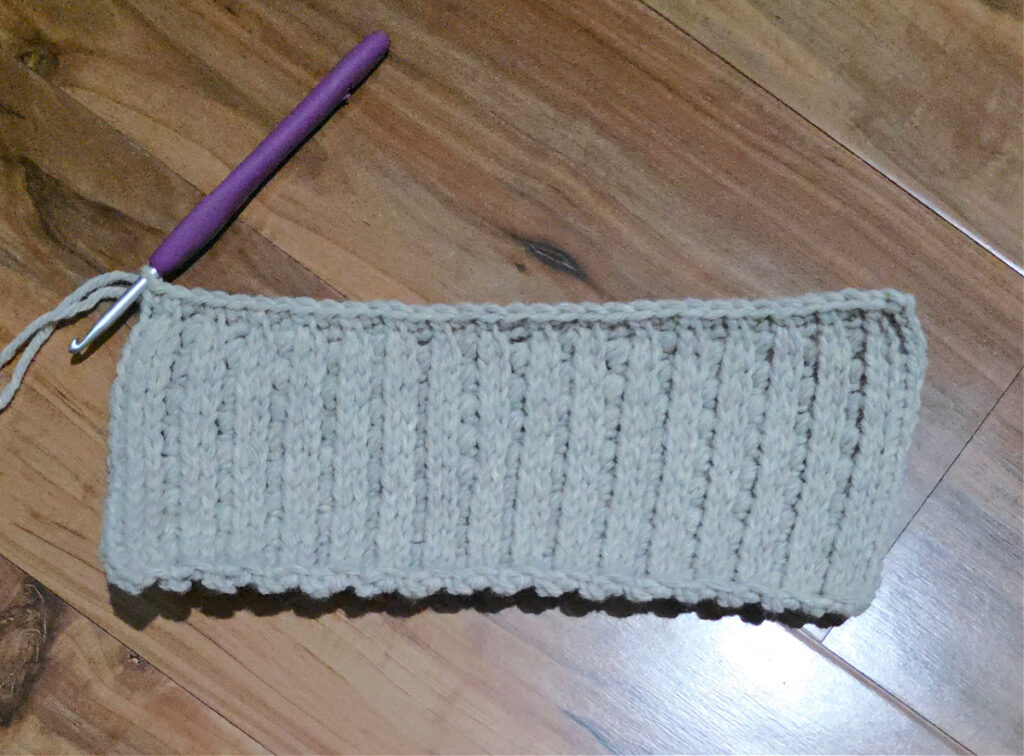 A grey crochet strip with a purple crochet hook against a wooden floor.