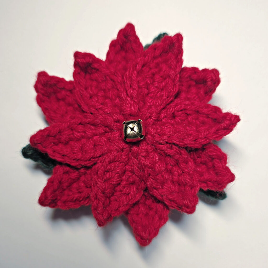 A completed red crochet poinsettia with green leaves and a small gold jingle bell in the center.