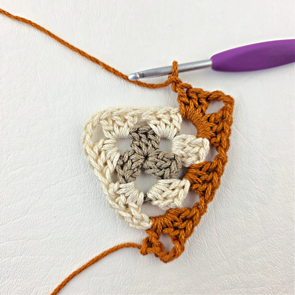 Round 3 of crochet granny stitch triangle with a purple crochet hook.