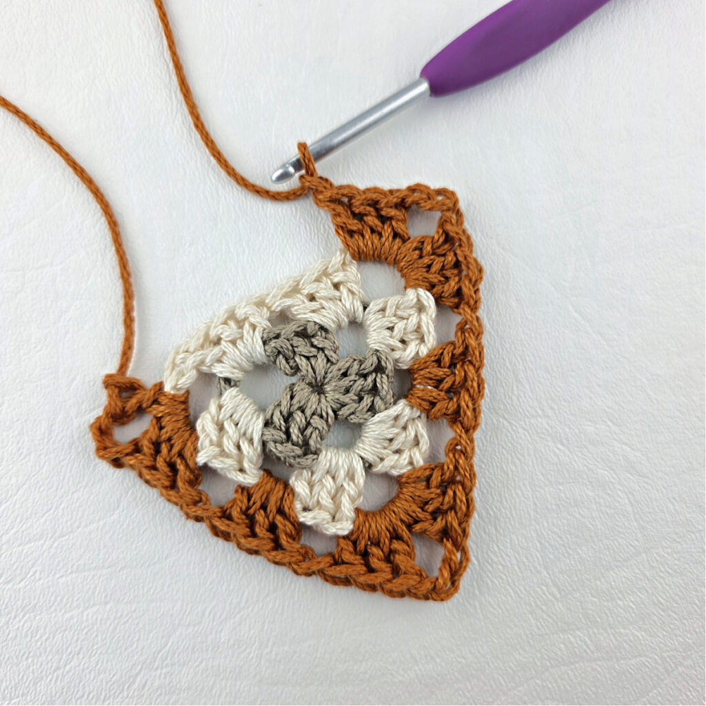 Round 3 of crochet granny stitch triangle with a purple crochet hook.
