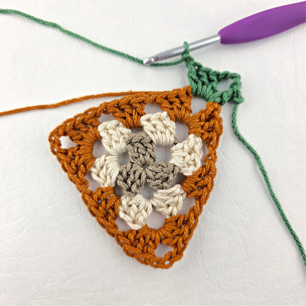 Round 4 of crochet granny stitch triangle with a purple crochet hook.
