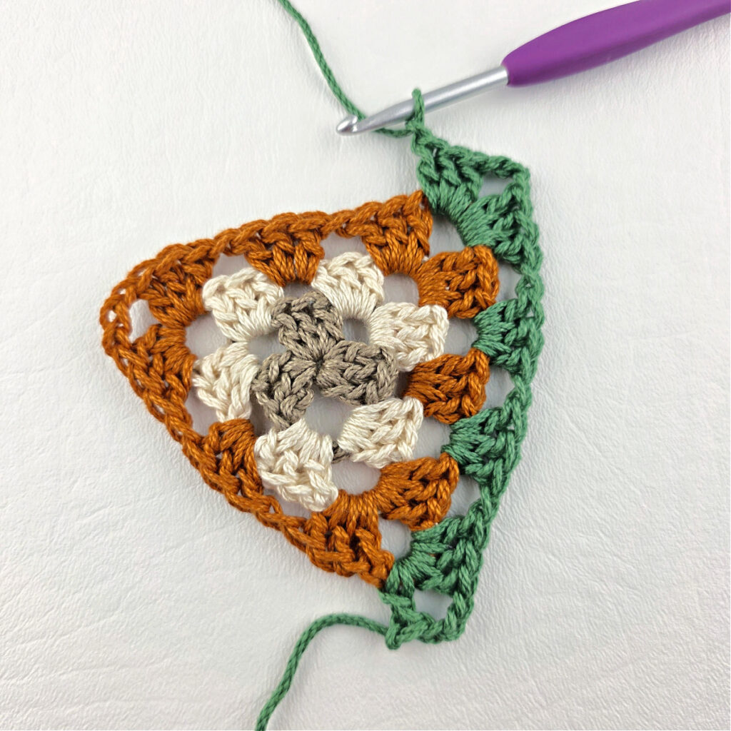 Round 4 of crochet granny stitch triangle with a purple crochet hook.