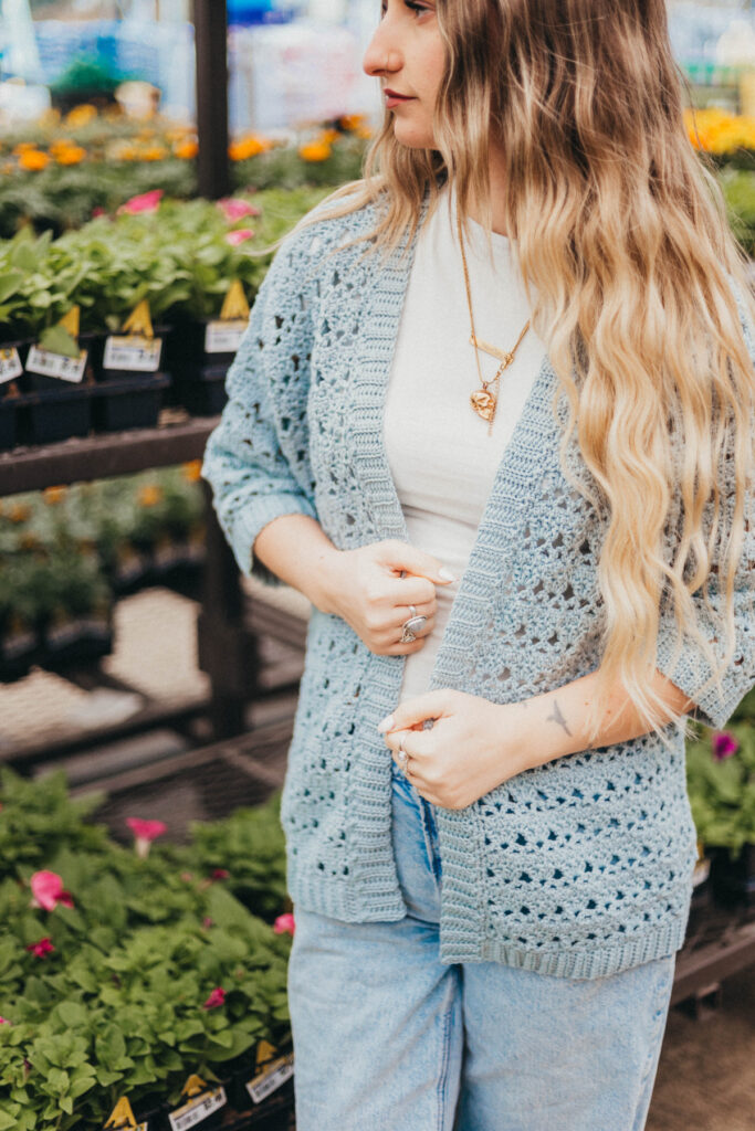 Girl wearing a light blue summer crochet cardigan and blue jeans with shelves of plants in the background.