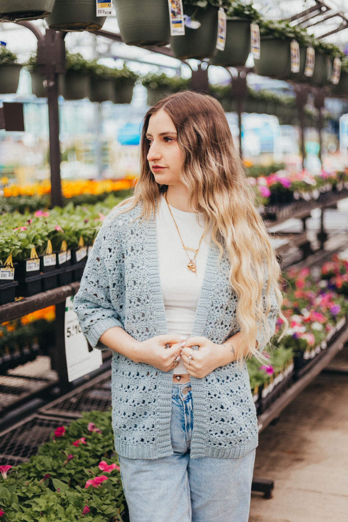 Girl wearing a light blue summer crochet cardigan and blue jeans with shelves of plants in the background.