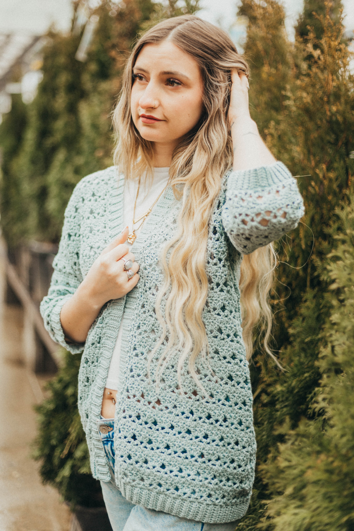 Girl wearing a light blue summer crochet cardigan and blue jeans with trees in the background.