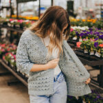 Girl wearing a light blue summer crochet cardigan and blue jeans with shelves of plants in the background.