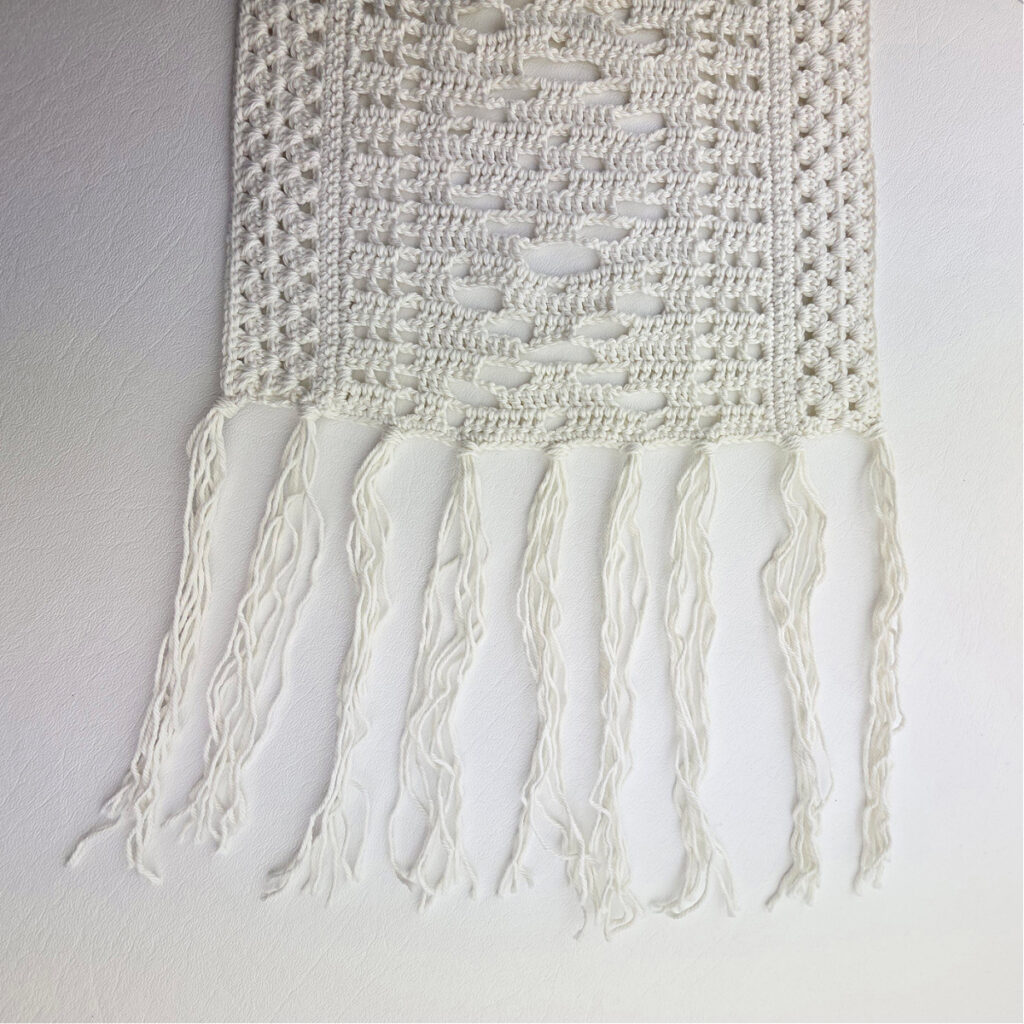 White lacy crochet table runner with a fringe against a white background.