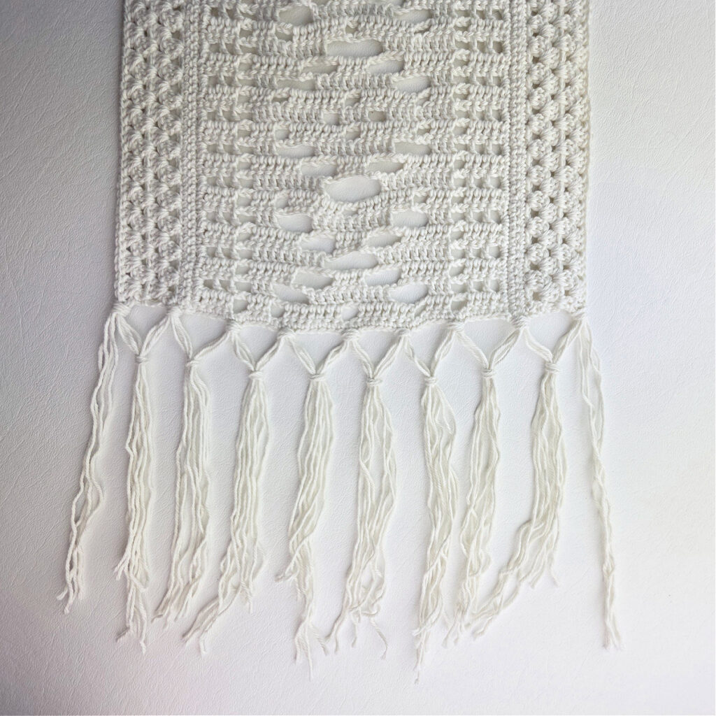 White lacy crochet table runner with a fringe against a white background.