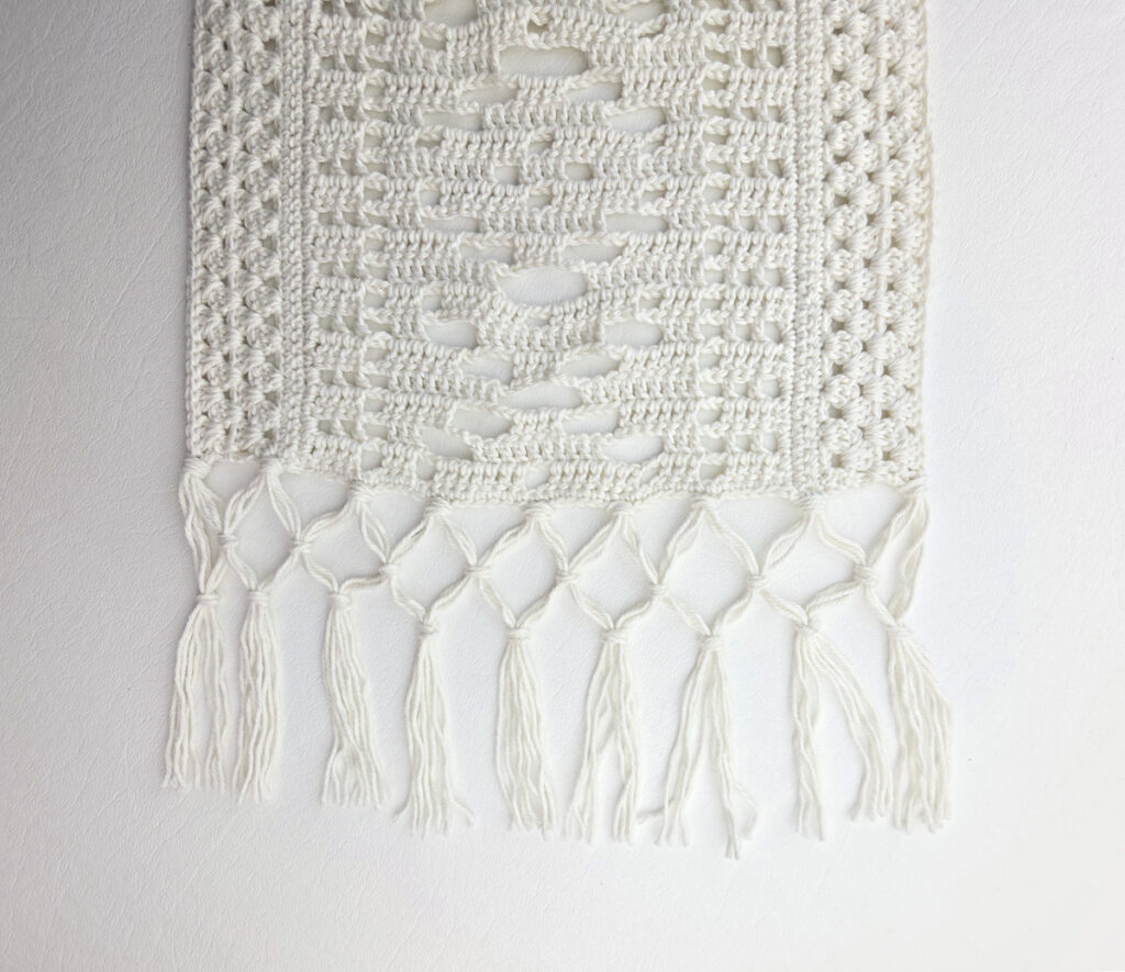 White lacy crochet table runner with a fringe against a white background.