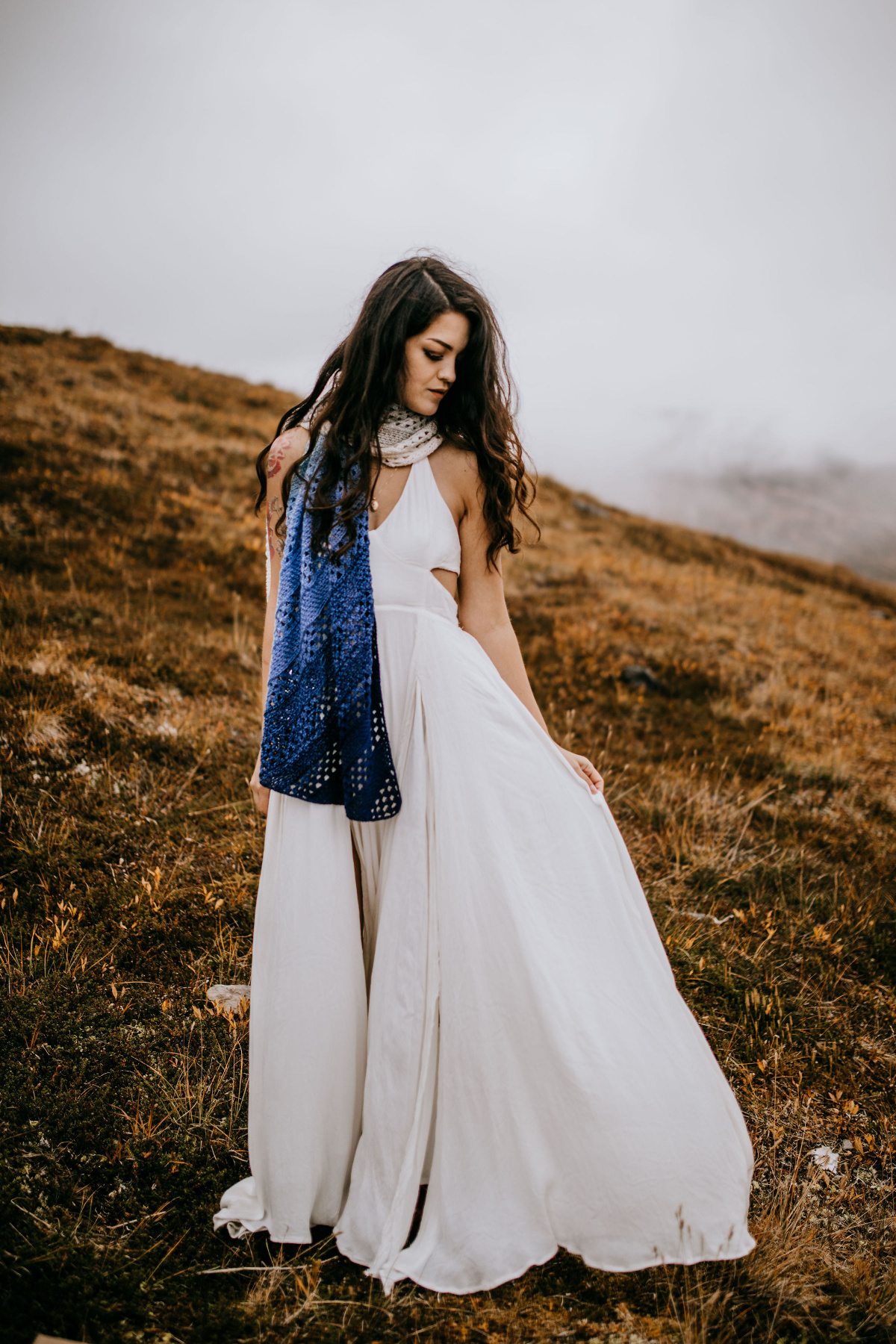 A woman dressed in a white dress and a crochet shawl standing on a hill.