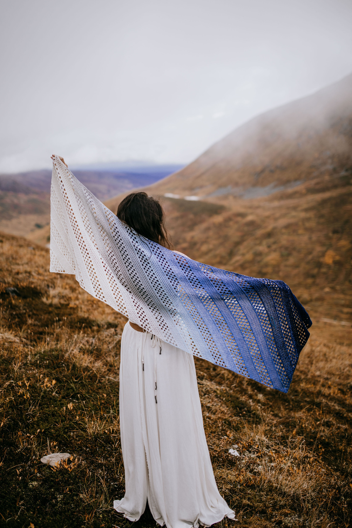 A woman dressed in a white dress and a crochet shawl standing on a hill.