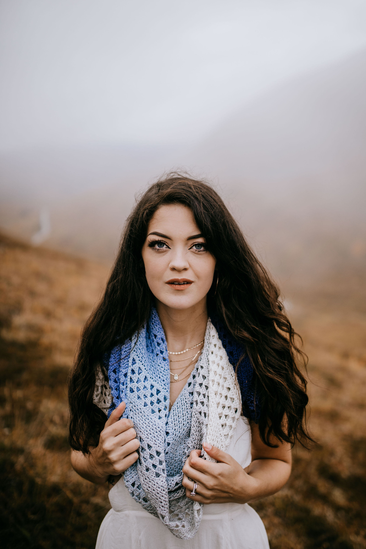 A woman dressed in a white dress and a crochet shawl around her neck standing on a hill.