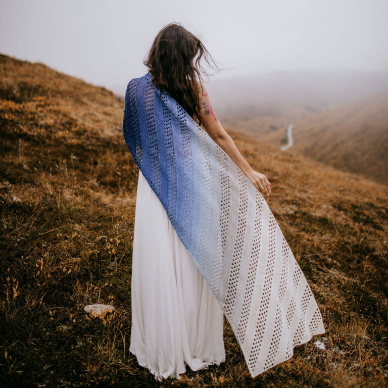 A woman dressed in a white dress and a crochet shawl standing on a hill with her back to the camera.